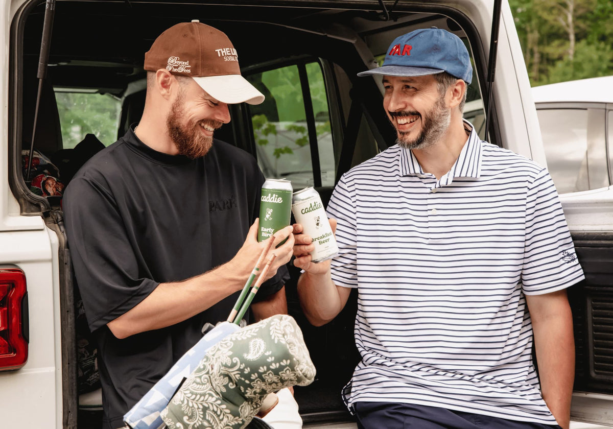 Two golfers laughing and cheersing cans of Caddie non-alcoholic beer at the back of a white SUV before a round of golf.